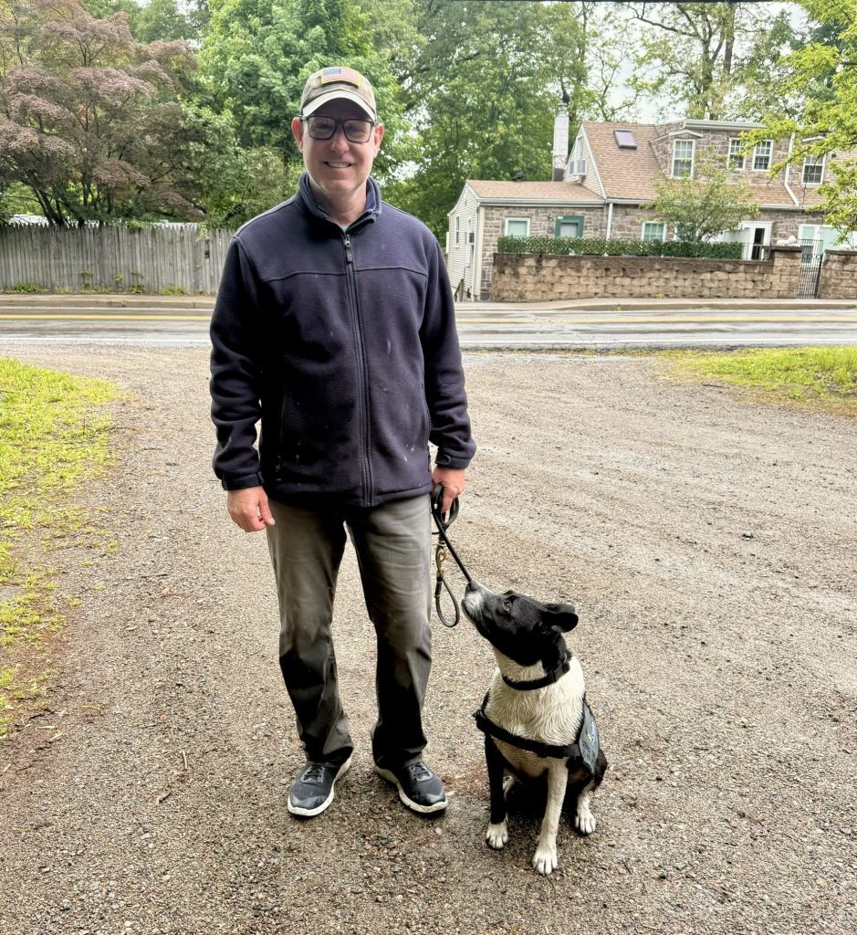 Smiling man in glasses, cap, and navy jacket stands on a gravel drive holding a leash as a black-and-white dog sits looking up at him; leafy suburban houses in the background.