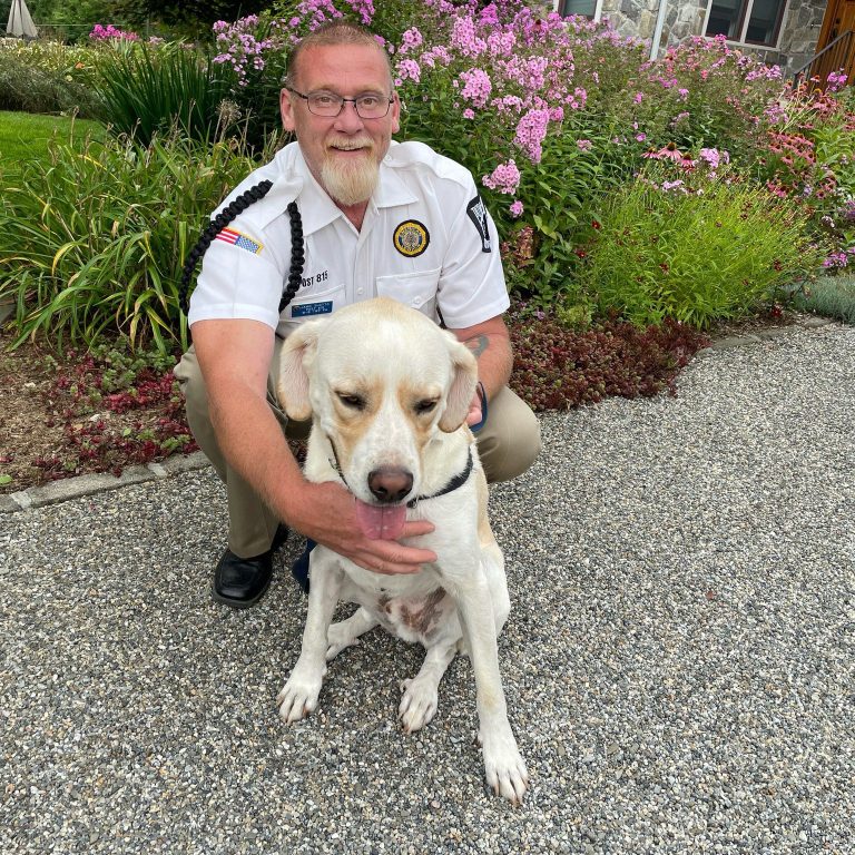 Smiling man in a white uniform shirt kneels on a gravel path, gently holding a light-colored Labrador mix; blooming garden and stone house in the background.
