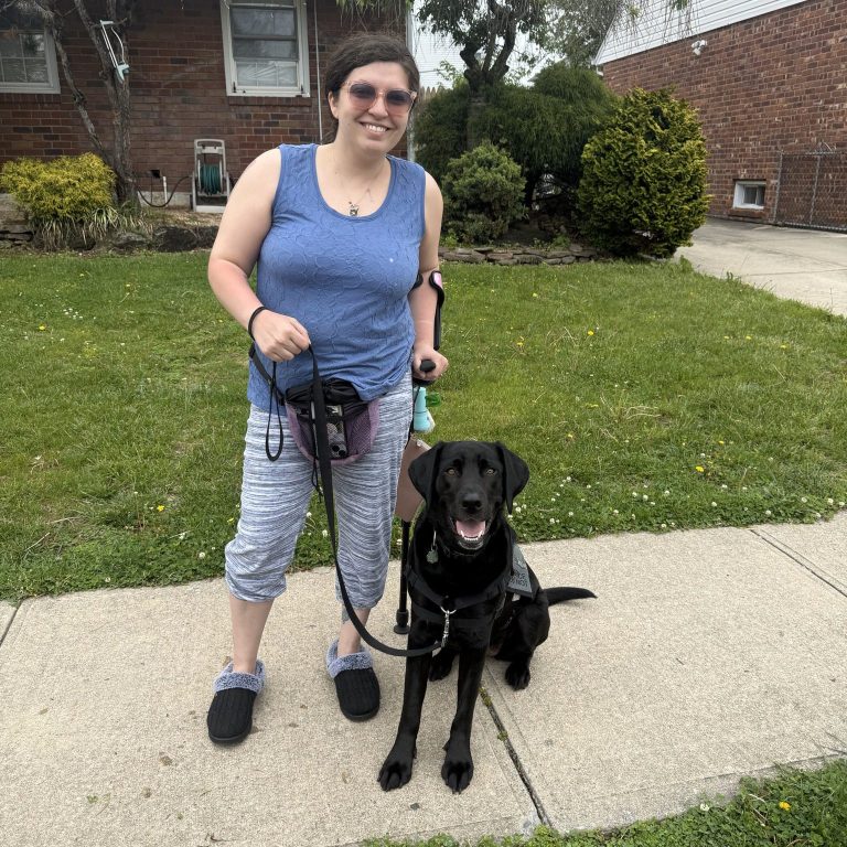 Woman with a forearm crutch stands on a sidewalk, smiling and holding the leash of a seated black service dog; suburban lawn and brick house behind.